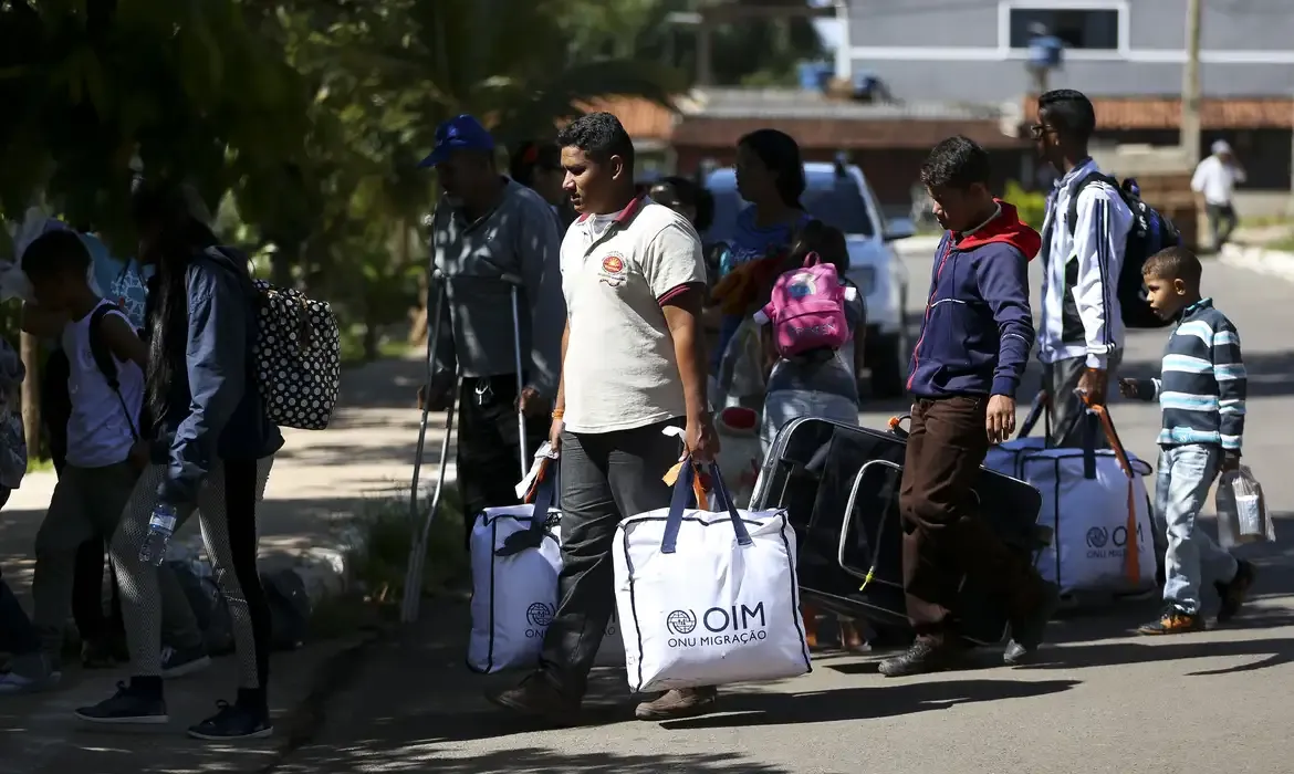 Imigrantes e suas famílias, com malas e bolsas, caminhando em direção ao apoio do governo brasileiro.